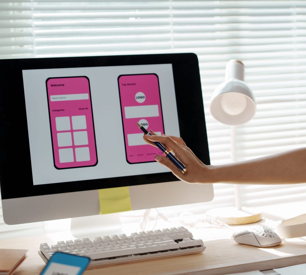 Designer's hand pointing at computer monitor developing new smartphone application or website design with pink UI elements